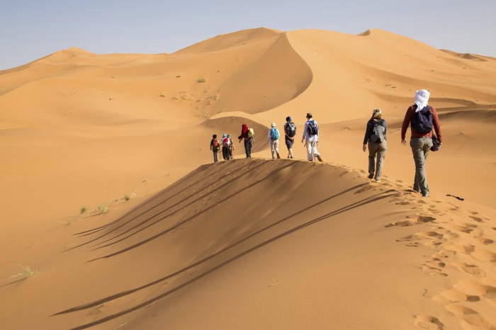 Group of people walking on sand dunes. Long shadows on the left side.